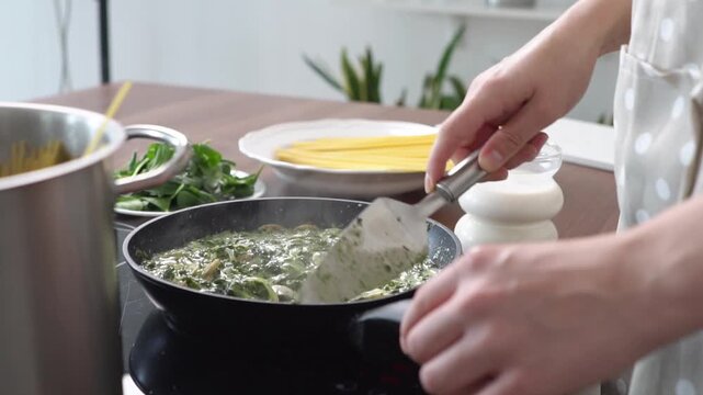 woman stir-fries mushrooms with spinach and sour cream in the kitchen, girl prepares pasta sauce in frying pan on the stove at home