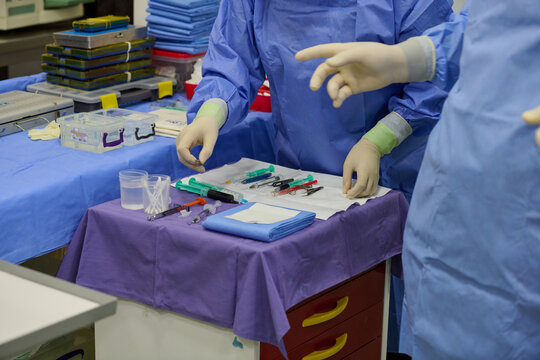 Surgeons in blue sterile gowns and latex gloves with green cuffs preparing instruments: syringes, scalpels, forceps on purple drape in operating room. Close-up sterile setup.