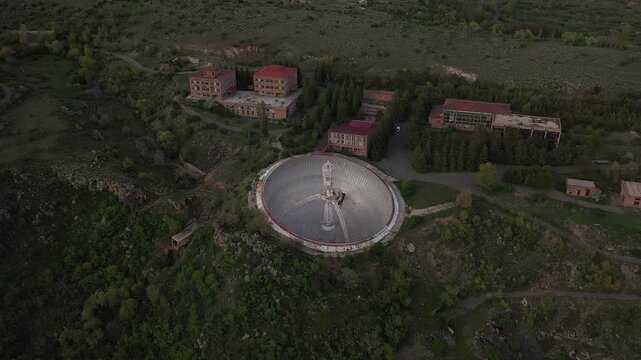  Aerial drone footage slowly zooms in on a massive abandoned radio telescope dish nestled within a mountainous landscape, revealing its intricate central structure and several nearby support buildings