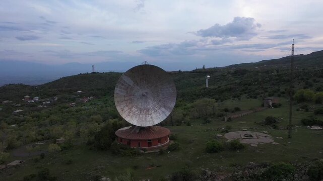Aerial drone video of a massive, abandoned radio telescope dish situated in a rugged mountainous landscape. The giant antenna overlooks a deep gorge under a cloudy evening sky.

