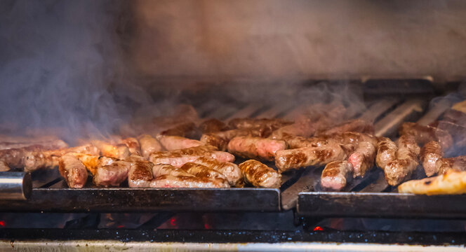Cevapi grilling on charcoal barbecue with smoke rising from hot grill, traditional Balkan street food preparation with minced meat sausages cooking over open fire.