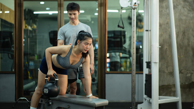 A woman is performing a kettlebell row on a bench while her trainer closely checks her form