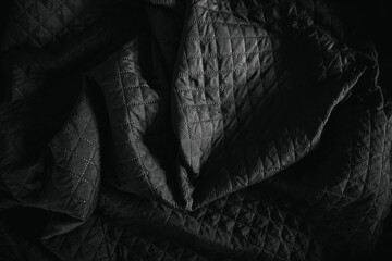 Fototapeta premium Close-up of black quilted bedsheet fabric in dramatic low light. Textured textile surface with deep shadows highlighting stitching pattern and soft folds.