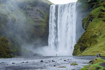 Obraz premium Huge waterfall surrounded by mossy rocky cliff and people walking towards it, Iceland