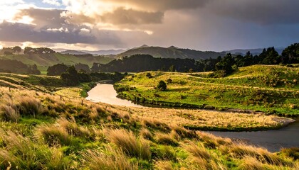 Obraz premium River winding through grassy hills, with sunlight breaking through clouds illuminating the landscape in golden hour