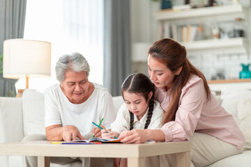 Fototapeta premium Asian grandmother and mother lean in together to help their young granddaughter with a coloring book at a living room table — a gentle three-generation learning moment.