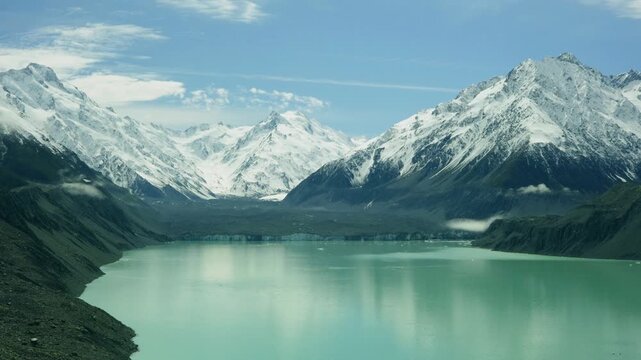 A pastoral telephoto view of a snow capped mountains above a Turquoise colored lake, with some beautiful light clouds and a little fog. a 4K video clip, mount Cook national park, New Zealand.