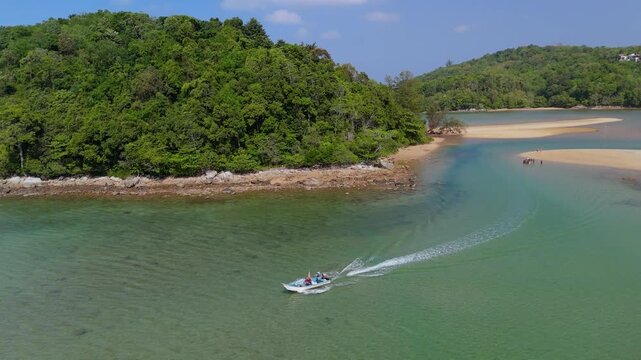 Motorboat moves swiftly across calm turquoise water, leaving a white wake. Green islands and sandy beaches define the tropical coast of Bang Tao Phuket Thailand