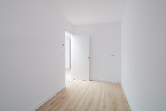 Empty room with white walls and wooden floor. A door is slightly open, leading to another space. Natural light illuminates the area.