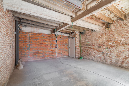 An empty room under renovation with exposed brick walls and concrete floor. The ceiling features wooden beams and electrical wiring is visible.