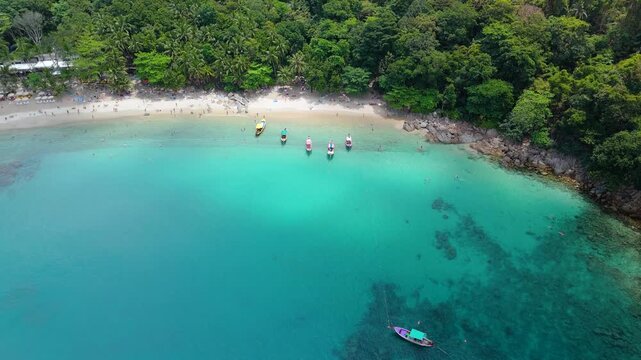 Banana Beach Phuket Thailand seen from above. Turquoise water holds longtail boats. Tourists swim and relax on the sand under green jungle trees