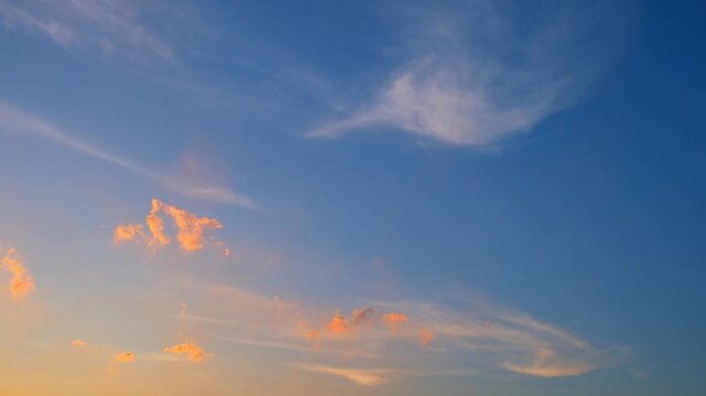 Sunset sky timelapse video capturing glowing cirrus cloud layers turning vivid pink and orange across the twilight sky, while small darker cloud patches drift beneath the colorful evening atmosphere.
