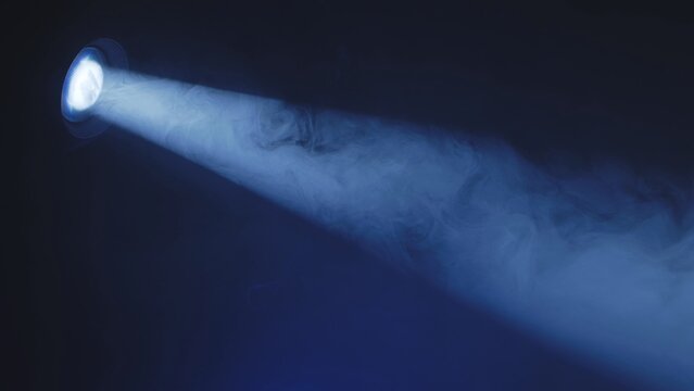Blue spotlight beam shining through dense smoke on a dark stage. Theatrical lighting effect.