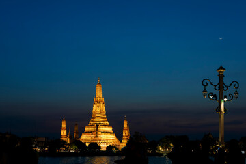 Naklejka premium Golden illuminated Wat Arun pagoda under a deep blue night sky with a crescent moon and ornate riverside lamp in Bangkok, Thailand.