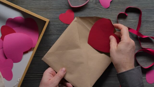A man's hand places a handmade Valentine's Day card into a brown kraft envelope on a rustic wooden table. Top view of a creative DIY process, preparing a romantic gift with craft paper and love