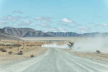 A wide gravel road winds through a vast, arid desert landscape towards distant mountains. A vehicle's dust trail lingers in the air under a clear blue sky. © marcodenaro