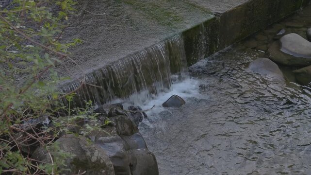 Water flows over a concrete berm into a rocky stream