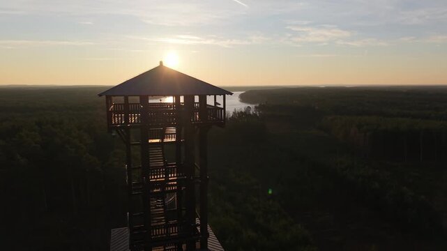 Aerial drone footage of a wooden observation tower surrounded by forest with a lake in the background at sunrise, warm morning light, peaceful nature landscape.