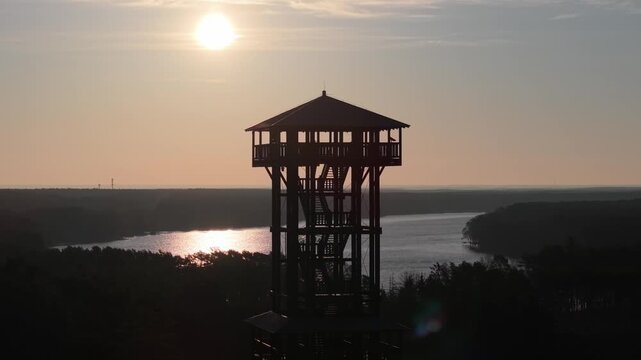 Aerial drone footage of a wooden observation tower surrounded by forest with a lake in the background at sunrise, warm morning light, peaceful nature landscape.
