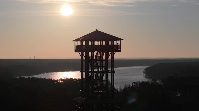 Aerial drone footage of a wooden observation tower surrounded by forest with a lake in the background at sunrise, warm morning light, peaceful nature landscape.