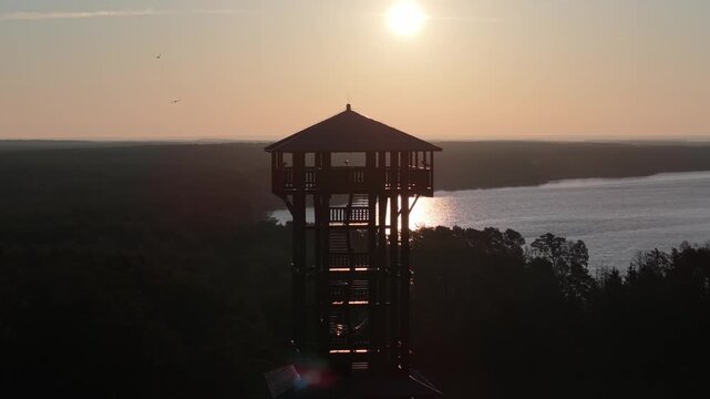 Aerial drone footage of a wooden observation tower surrounded by forest with a lake in the background at sunrise, warm morning light, peaceful nature landscape.