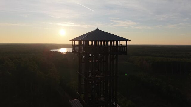 Aerial drone footage of a wooden observation tower surrounded by forest with a lake in the background at sunrise, warm morning light, peaceful nature landscape.
