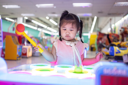 Adorable Little Girl Playing Whack-A-Mole Arcade Game with Toy Hammer in Game Center,A cute Asian toddler girl focuses intently on playing a classic whack-a-mole redemption game at an indoor amusement