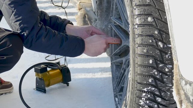 Man inflating car tire with portable electric compressor on snowy road. Driver checking tire pressure in wintertime. Vehicle maintenance and emergency roadside assistance during cold weather.