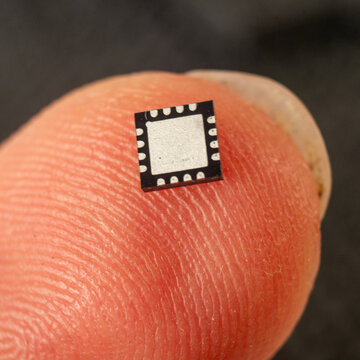 Bottom/pad side facing up, dark background
QFN microchip resting on a fingertip showing the silver exposed pad and peripheral contacts, warm skin tones contrasting with the dark background, emphasizin
