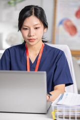Fototapeta na wymiar Young Asian female nurse wearing navy blue scrub looking heavily focused and serious while typing on silver laptop computer sitting at modern medical clinic desk reviewing patient health record
