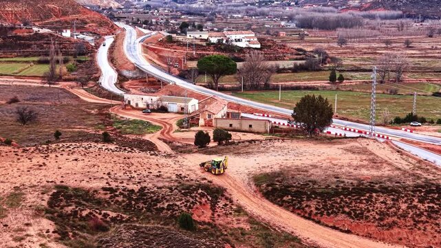 Aerial view of backhoe loader near rural road bridge. Wide landscape featuring construction machinery, red soil fields, a concrete bridge, and farm buildings under daylight.