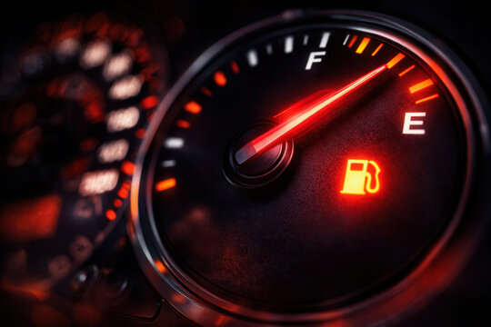 Closeup of a car dashboard fuel gauge showing empty with a glowing low fuel warning icon. Dramatic lighting highlights the urgency of running out of gasoline while driving.