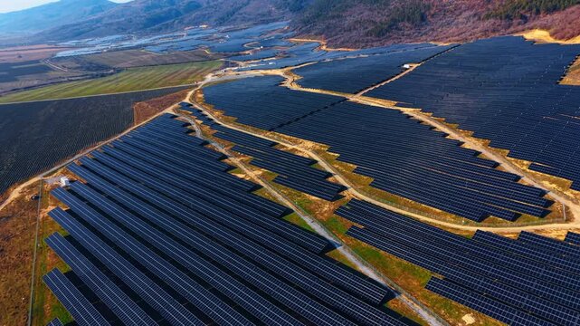 High altitude aerial view of massive solar power complex. Drone shot of vast solar panel fields integrated into hilly rural landscape under clear sky.