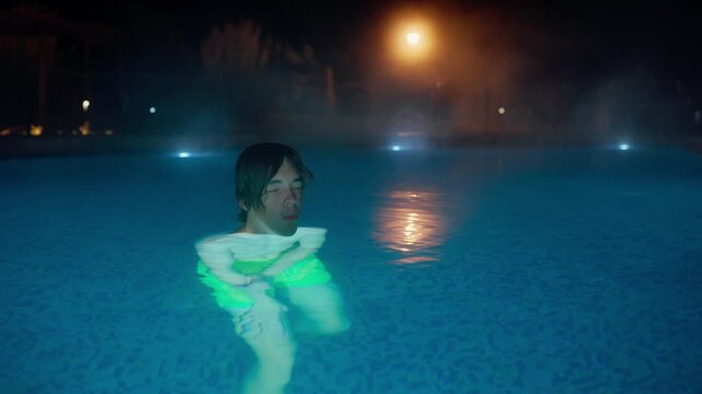 A young man with closed eyes relaxes in a steamy outdoor swimming pool at night. The warm water, mist, and soft lighting create a tranquil, meditative atmosphere for wellness and escape