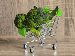 Fresh green broccoli florets placed inside a miniature toy shopping cart on a wooden surface, representing healthy food choices. © Mariia135