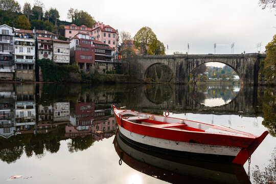A picturesque view of the historic S&atilde;o Gon&ccedil;alo Bridge and the T&acirc;mega River in Amarante, Portugal. In the foreground, a traditional red and white wooden boat floats calmly on the water