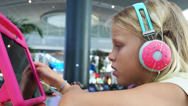Young child wearing colorful donut shaped headphones focuses on a pink tablet screen while waiting in a modern airport terminal lounge