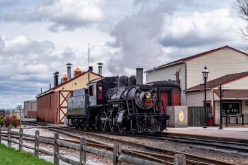 A steam locomotive is at a train station with smoke rising from its chimney. The train is near a wooden fence and old buildings under a cloudy sky. People wait nearby as the train arrives.