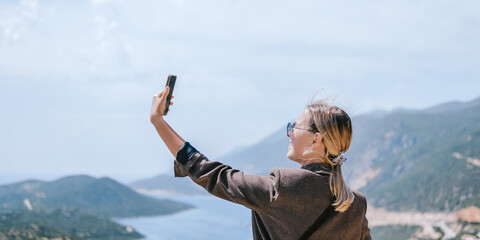 Travel blogger filming live stream on smartphone against mountain coastline, Gen Z female tourist on viewpoint, panoramic sea and hills, social media content creator, sunny outdoor vacation vibe. © shintartanya