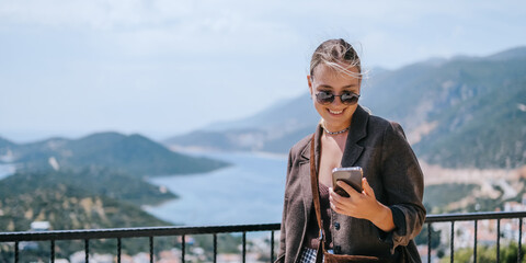 Travel blogger filming live stream on smartphone against mountain coastline, Gen Z female tourist on viewpoint, panoramic sea and hills, social media content creator, sunny outdoor vacation vibe. © shintartanya