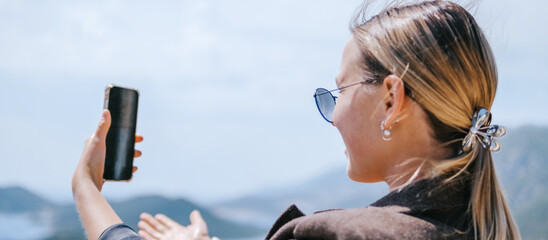 Travel blogger filming live stream on smartphone against mountain coastline, Gen Z female tourist on viewpoint, panoramic sea and hills, social media content creator, sunny outdoor vacation vibe. © shintartanya