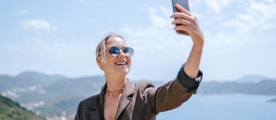 Travel blogger filming live stream on smartphone against mountain coastline, Gen Z female tourist on viewpoint, panoramic sea and hills, social media content creator, sunny outdoor vacation vibe. © shintartanya