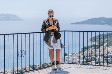 Travel blogger filming live stream on smartphone against mountain coastline, Gen Z female tourist on viewpoint, panoramic sea and hills, social media content creator, sunny outdoor vacation vibe. © shintartanya