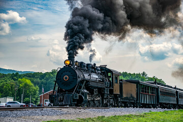 A steam locomotive pulls away from a train station. Dark smoke rises from the engine while freight cars travel behind. Cars are parked nearby.