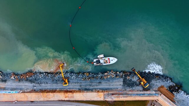 Top down view of coastal dredging with two excavators. Industrial work on rocky shore with boat and sediment control curtain.