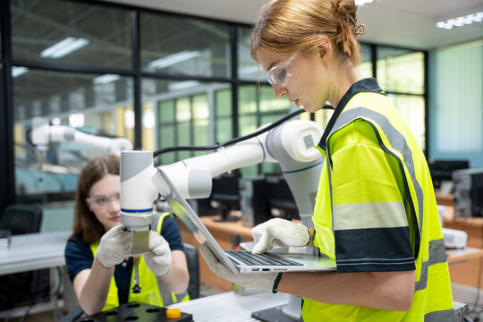 Professional female engineers using laptop to program and control collaborative robotic arm in modern industrial laboratory, focusing on smart factory automation and future robotics technology.