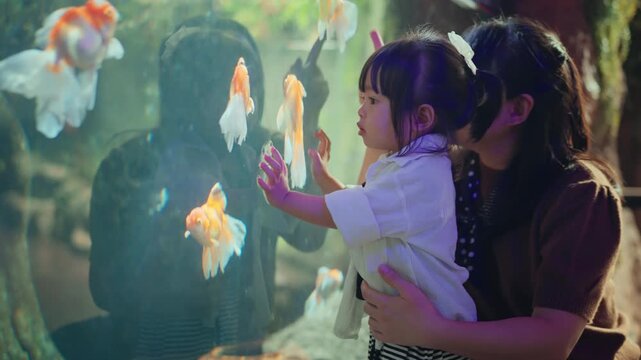Curious little Asian girl looking at goldfish in aquarium while her mother points at the fish to teach her. Happy family bonding and learning about aquatic life together, showcasing childhood wonder