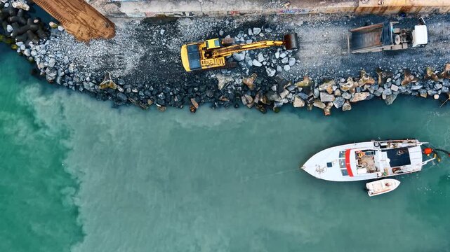 Aerial view of coastal dredging and shore reinforcement works. Top down perspective of stationary excavator near truck and water edge.