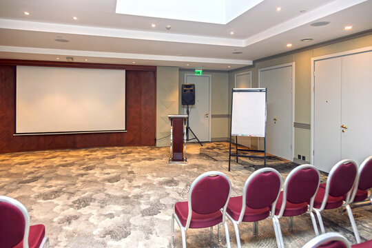 Empty corporate conference room with large projection screen, flipchart and rows of red chairs set up for a business presentation, seminar or training event in a modern meeting space.