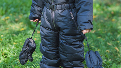 Child holding mittens, wearing winter snowsuit, standing outdoors in green grass